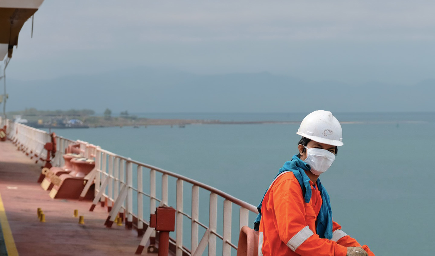 A masked seafarer sits on the side of a large vessel.