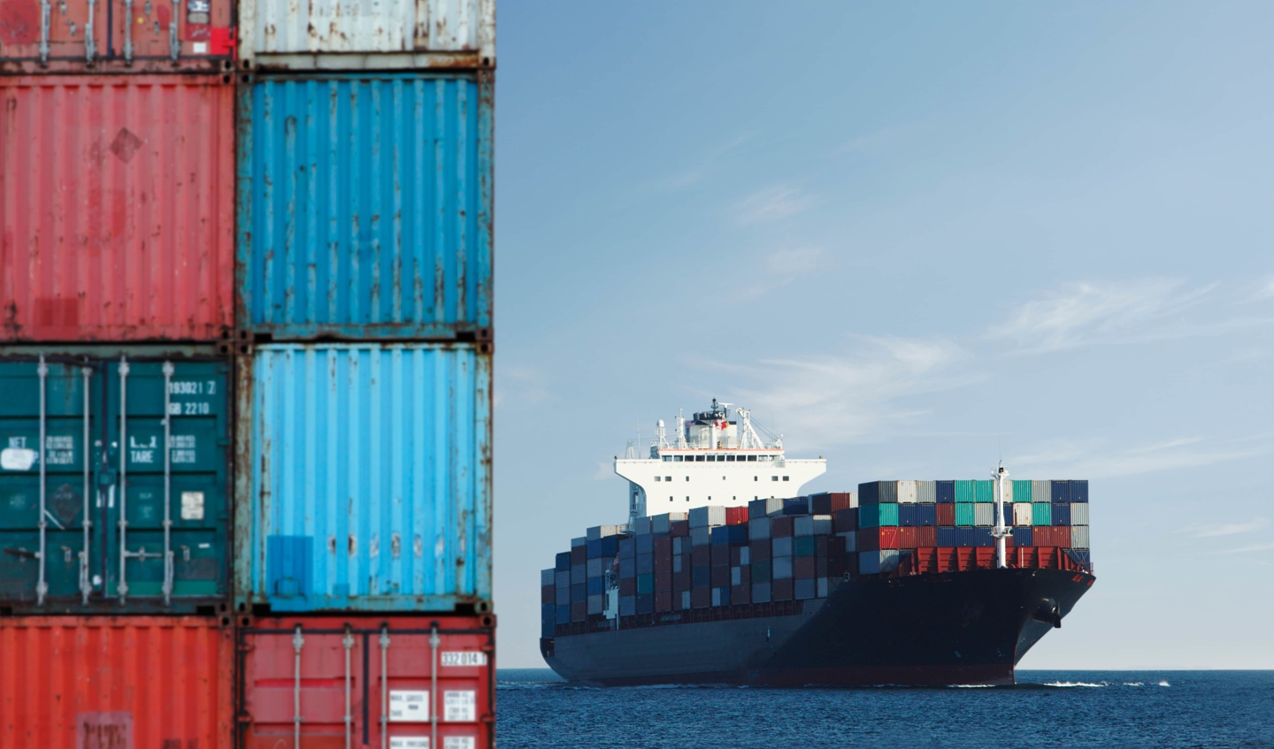 A shipping container sits in the foreground and a ship on the sea in the background.