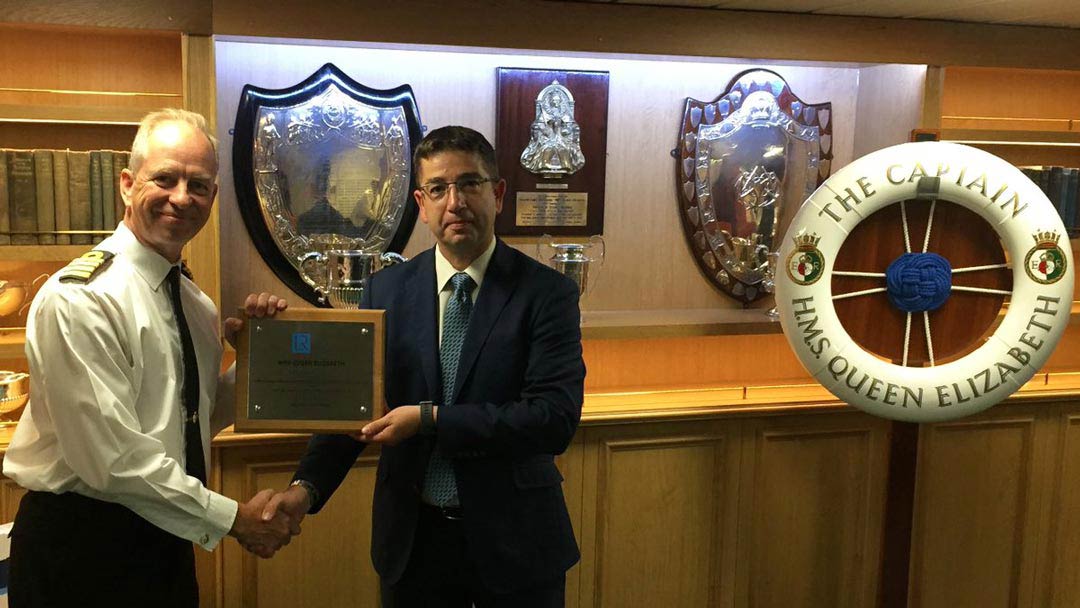 Image of class certificates being presented to the ship’s Commanding Officer by Captain Kyd in front of trophy lined wall