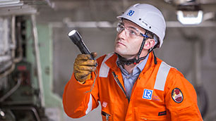 Loyd's Register Surveyor in orange overalls with helmet on and torch examining structure