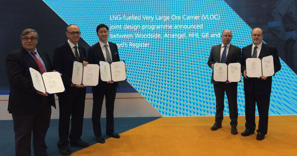 Five executives standing holding framed documents in front of bright blue graphic backdrop with design programme text.