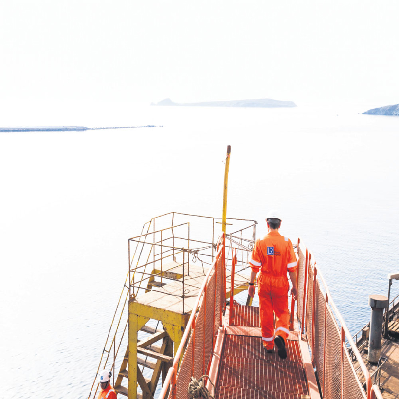 Seafarers at work on scaffolding, ocean and mountains in the background.