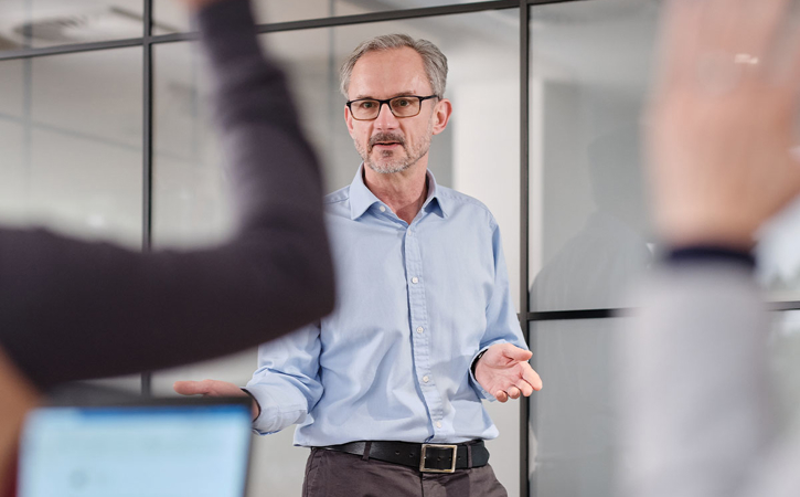 A trainer standing at the front of a room, framed between blurred hands to show they are answering questions.